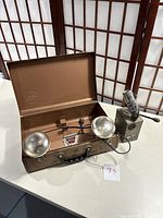 Photo showing the two vintage remote flash units on a white surface with one unit open in brown metal case displaying dual reflectors and attached cords.