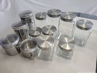 Full set of 12 storage jars arranged on white surface: 8 glass jars with stainless steel lids and 4 metal jars with snap lids.