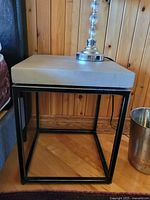 Single side table photographed with a lamp on top, showing the square grey faux concrete surface and black open cube metal base.