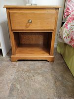 Front view of one solid oak bedside table showing drawer with faceted glass knob and open shelf below, standing on tiled floor next to bed.