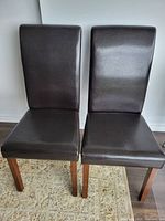 Two brown faux leather parsons style chairs shown side by side on a rug with wooden flooring. The chairs have tall backrests with a rolled top, and wooden legs.