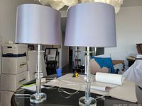 Pair of side table lamps with clear glass bases and silvery grey shades shown on a glass table with background of office items.