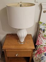 Full view of one white table lamp placed on a wooden nightstand beside a bed with floral bedding.