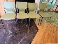 Four green dining chairs lined up on a dark marble floor. Upholstered seats and backs appear stiff, likely faux leather. Chrome legs and frame visible.