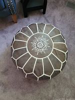 Top view of round blue leather pouf showing embroidered white floral pattern and stitched segments on top surface.