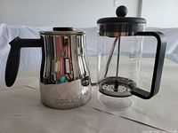 Photo shows both the Bodum Bistro gooseneck kettle and the Bodum French Press coffee maker side by side on a white surface, highlighting the polished stainless steel finish of the kettle and the clear glass of the French press.