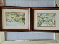 Two framed watercolor paintings showing Cotswold scenes, laid flat on a surface. Both artworks have wooden frames with matting and gold trim around the image area.
