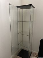 Glass display cabinet door open showing interior glass shelves and black/brown veneer top and base.