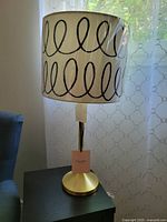 Photo showing entire lamp with white shade featuring black looping scribble pattern. Gold toned base and tag visible.