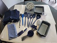 Overview photo showing navy towels, blue and white dishcloths, silicone utensils, measuring cups and spoons, can opener, spatulas, tongs, strainers, loaf pans, square pan, cookie sheets, and measuring container on table.