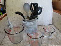 Three clear glass Fire King measuring cups (1, 2, 4 cup sizes) with red measurement markings, a gray metal utensil holder, and six black kitchen utensils with wooden handles shown on a beige fabric chair.