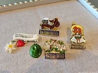 Four colorful metal lapel pins themed around Kitchener-Waterloo Oktoberfest arranged on a neutral fabric background. Pins include a beer stein with dachshund, Oktoberfest character, green alpine hat, and Edelweiss flower with heart and festival text.