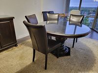 Round solid wood pedestal dining table with six dark brown leather chairs around it, situated on a patterned carpet near a window with city view.