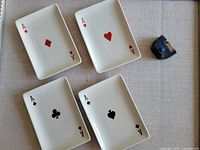 Four rectangular white ceramic plates each emblazoned with a different Ace symbol: diamonds, hearts, clubs, spades. A small blue dice with visible wear sits beside the plates on a fabric surface.