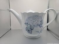 Front side view of the small white ceramic watering can featuring the hydrangea floral pattern and curved handle.
