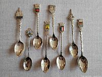 Full view of 7 souvenir spoons laid out on fabric background, showing various French landmarks and crests on the spoon tops and intricate twisted handles.