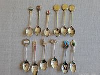 Full view of 11 souvenir spoons arranged on fabric background, showing variety of crest and castle designs on handles.
