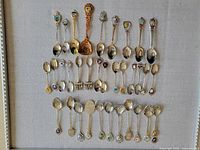 Overview photo showing thirty souvenir spoons arranged in rows on light gray fabric; spoons vary in size, handle design, and decorative emblems.