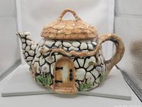 Front angle showing thatched roof lid, stone wall texture, and wooden door detail on teapot shaped like cottage.