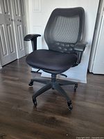 Front and side angle view of black SXO office chair showing seat, armrests, mesh backrest, and caster wheels on a wood floor.
