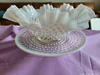 Vintage opalescent hobnail glass bowl placed on hobnail glass plate on lavender fabric background. Bowl has fluted edges with opalescent white and clear base, and rounded hobnail texture all over body.