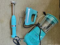 Three turquoise kitchen appliances on carpet: Kitchenaid immersion blender with whisk attachments, hand mixer; Cuisinart blender with control panel visible