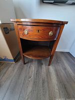 Front view of mahogany bowfront bedside table with one drawer and one shelf, brass circular handles and keyhole visible.
