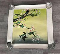 Full view of the square Chinese watercolor painting on silk showing two birds with blossoming branches, mounted with a white fabric border and held by glass weights at corners.