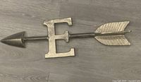 Photo of silver metal arrow wall hanging featuring a large letter 'E',' with arrowhead on one side and fletching on the other, placed on wood floor.