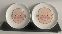 Two white ceramic plates side by side, each with a cartoon-style face in the center. The faces have rosy cheeks and simple features. Displayed on a black shelf against a white wall.