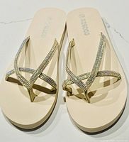 Top view of beige flip flops showing soft sole and rhinestone-studded thong straps crossed on top.
