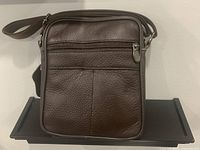 Front view of dark brown genuine leather men's crossbody bag on a black shelf against a beige wall, showing one zippered pocket on the front.