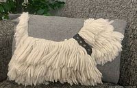 Front view of shaggy dog pillow showing yarn tassels, black studded collar, and grey background fabric on couch.