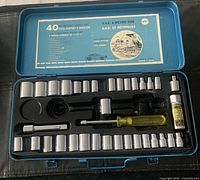 Metal blue case open displaying a 40-piece socket set with sockets arranged in rows, ratchet handle, extensions, screwdriver handle, and bottle of lubricant. Label with specifications attached to the lid.