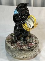 Front view of black cat figurine wearing blue scarf holding yellow basket with white cloth on wood plank styled round base