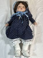 Full body shot of porcelain doll in blue dress with white lace trim, white lace socks, and white shoes, with brown hair and blue bonnet.
