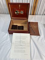 Open cigar humidor showing interior with built-in round hygrometer and humidifier, two brown textured cigar holders inside, humidifier brush attached to lid, instruction booklet and printed instruction sheet laid out beside it.