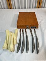 Top view of 5 curved stainless steel knives with hammered texture handles, arranged side by side on a table next to yellow cleaning cloths. Wooden box is closed.