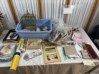 Overview of craft supplies and decorative items laid on table including birdcage, storage bins, frames, and bags of materials.