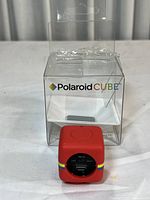 Red Polaroid Cube camera placed next to its clear plastic packaging box, showing branding and compact cube shape.