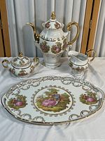 Full display of the porcelain tea set including teapot, sugar bowl, creamer, and serving tray with Victorian scenes and gold accents