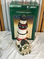 Photo showing the lighthouse figurine next to its original green box marked 'Santa's Workbench Collection Victorian Series'. The figurine includes an electric cord and is 12 inches tall.