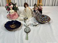 Overview of the entire vintage doll and collectible figurine assortment laid out on table with various dolls, porcelain figurines, boxes, hand mirror and decorative dish.