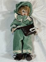 Full view of the porcelain doll lying on fabric background showing the green velvet outfit with white lace, brown bag, and black shoes.