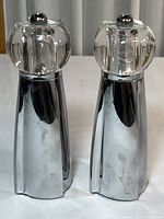 Full view of two metal and clear acrylic salt and pepper grinders showing silver metal bodies and faceted clear acrylic tops with metal knobs