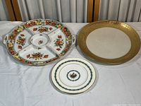 Three porcelain plates displayed on table: large divided floral with handles, white plate with gold rim, small plate with blue floral wreath and bird.