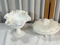 Two white milk glass compotes on a table, one tall with ruffled edge and pedestal base, one shorter covered dish with hobnail lid and feet.