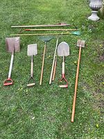 Photo showing 7 long-handled garden tools including shovels, rakes, and a heavy tamping tool laid out on grass.
