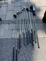 Top-down angled photo showing the golf clubs arranged on a paving surface; multiple clubs including drivers, irons, putters arranged in a row with visible grips and shafts.