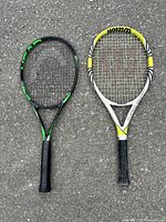 Two adult tennis rackets laid side by side on outdoor pavement, Wilson THREE.SEVEN white/yellow and HEAD Metallix black/green, showing front side with strings and grips visible